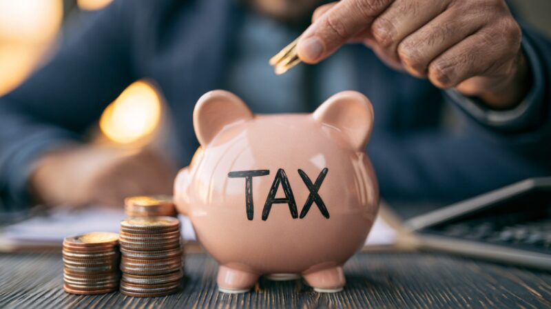 Close-up of a hand placing coins into a piggy bank labeled tax, with stacked coins on the table.