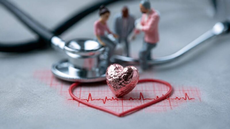 Heart symbol with ECG line, stethoscope and blurred medical figures in the background.